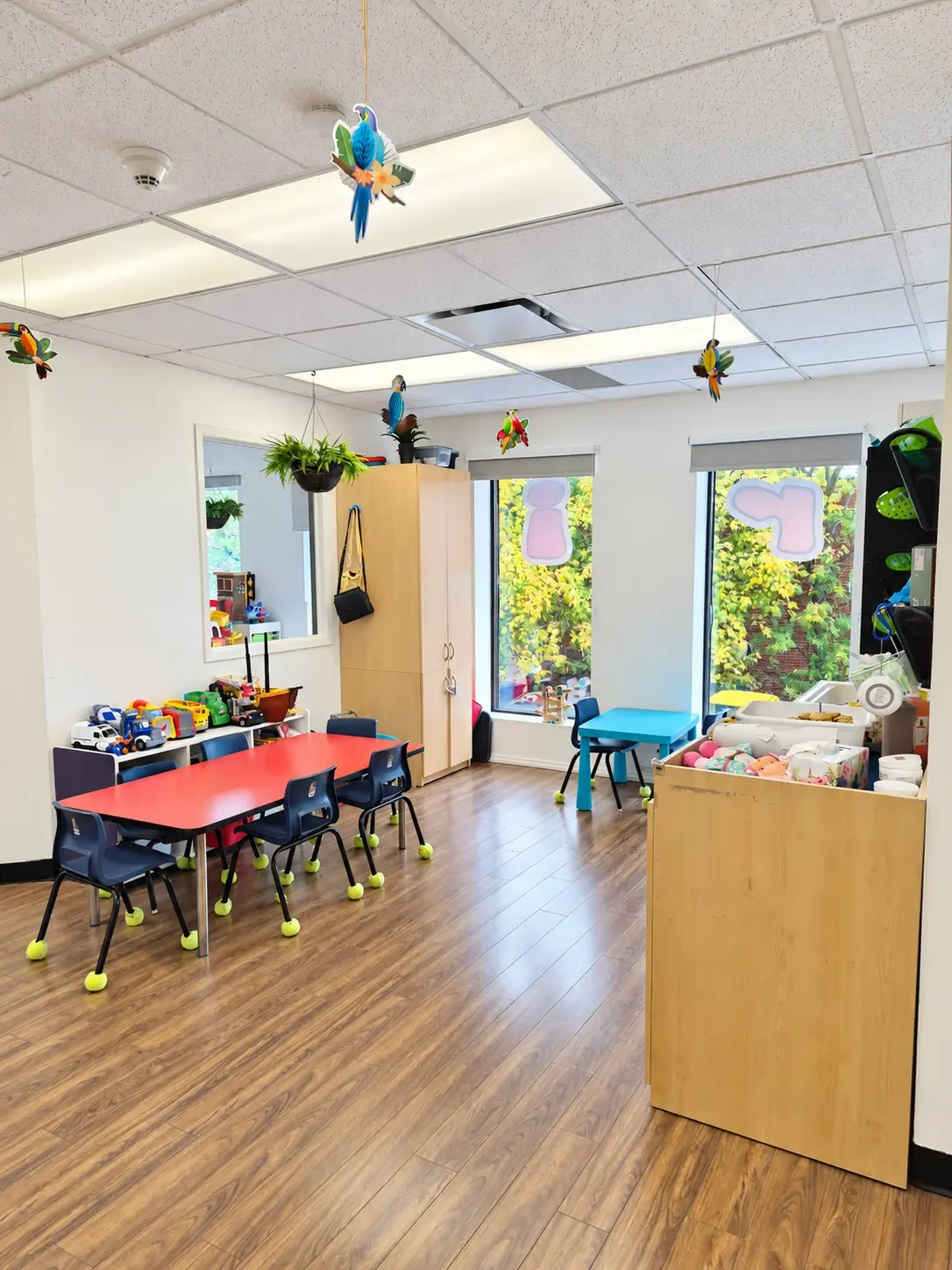 Salle avec une table rouge entourée de petites chaises, des jouets sur une étagère et deux grandes fenêtres donnant sur des arbres