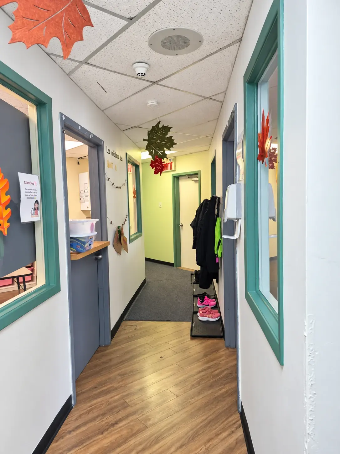 Corridor aux murs clairs avec fenêtres intérieures donnant sur les salles, crochets à manteaux et tapis d’entrée